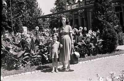 Woman and young boy pose near a grand villa, likely lakeside. The woman carries a picnic basket, showcasing mid-century fashi...