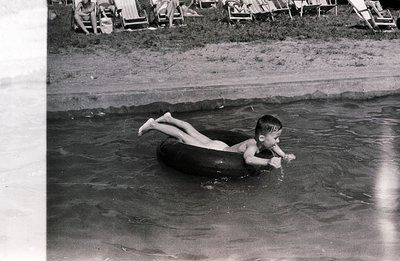 A young boy floats on a black inner tube in calm water. Background shows a beach with multiple lounge chairs. Appears to be a...