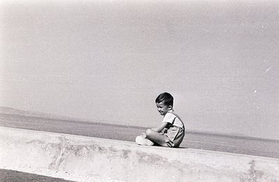 A young boy sits on a concrete retaining wall overlooking the sea. He wears short shorts and a short-sleeved shirt, appearing...
