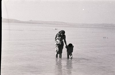 A woman and young child are seen wading in shallow water, possibly a sea or lake. The scene appears calm, with distant hills ...