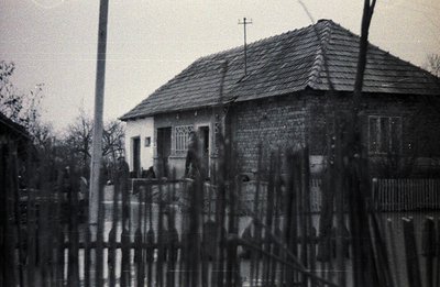 A modest, single-story house with a steeply pitched, shingled roof is framed by a weathered wooden fence. Utility pole and ba...