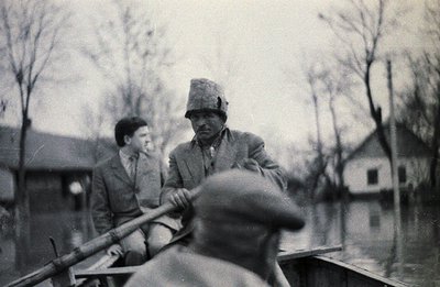A man rows a small boat carrying a young boy through floodwaters, likely post-war period. Sparse trees and a simple building ...