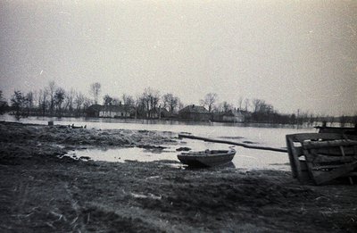Flooded landscape with a small rowboat beached on muddy ground. Several simple, wood-frame houses are visible in the distance...
