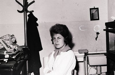 A young woman sits in what appears to be a small, utilitarian room, possibly a caretaker's office. She wears a crisp white bl...