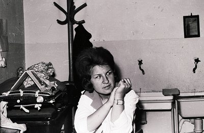 A young woman with a short, styled hairstyle sits thoughtfully. She wears a white blouse & wristwatch. A dark coat hangs on a...
