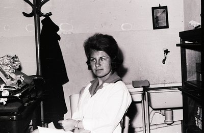 A young woman in a crisp white uniform sits in a simple chair, posed near a washbasin and shelving unit. The photograph, like...