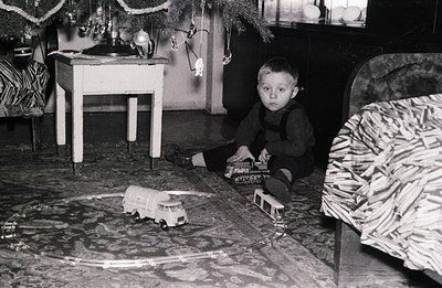 A young boy, wearing overalls, sits cross-legged amidst a miniature train set on an ornate rug. Decorated with sparse ornamen...