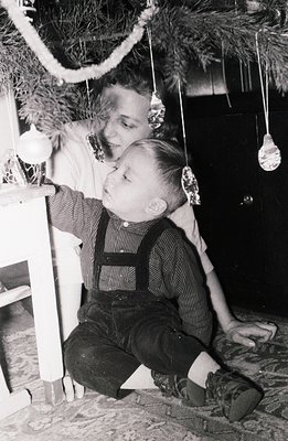 A young boy in overalls sits near a decorated Christmas tree, partially obscured by a smiling adult. Simple ornaments hang fr...