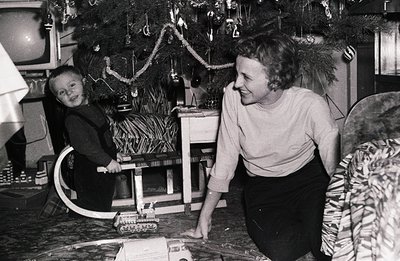 A young boy and woman, likely mother and son, interact near a decorated Christmas tree. The boy playfully poses with a toy tr...