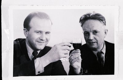 Two formally attired men in suits and ties clink wine glasses in a gesture of celebration. The photo, likely a candid snapsho...
