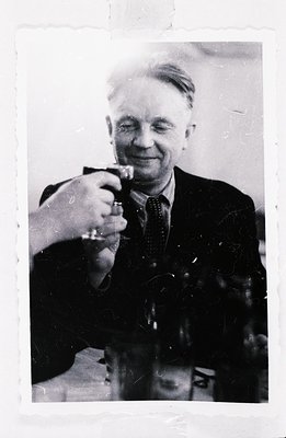 A monochrome image shows a smiling man raising a glass in a toast. He wears a dark suit and patterned tie, appearing to be in...