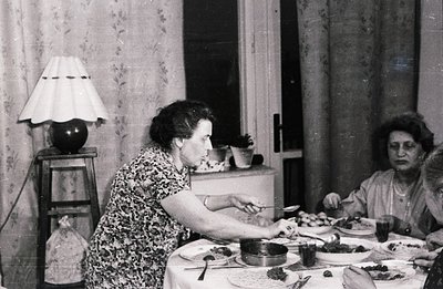 A family gathers for a formal indoor meal. A woman serves food from a pot with a spoon. Visible are plates, silverware, and a...