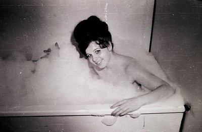 A woman with a stylized beehive hairstyle soaks in a frothy bathtub. She gazes directly at the camera with a slight smile. Th...