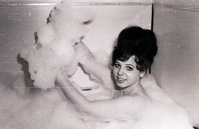 A woman with a beehive hairstyle is submerged in a large bubble bath, playfully gesturing toward the camera. The image, likel...