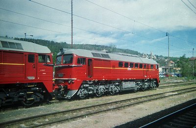 Two red diesel-electric locomotives sit on a railway line, likely in a depot or siding. The locomotives exhibit a boxy, strea...