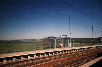 A solitary figure stands on the platform of the Černovice train station, framed by a simple, concrete shelter. Rolling hills ...