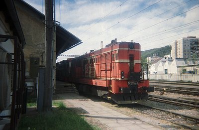 A red diesel locomotive (number 742 516-8) stands on tracks at a railway station platform, framed by a weathered station shel...