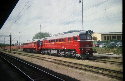 A red electric locomotive, likely a Bulgarian State Railways model, speeds along tracks under power lines. The streamlined de...