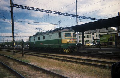 A green electric locomotive, likely Bulgarian State Railways (BDZ) type Е669, sits on tracks near a station platform. Visible...