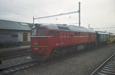 A red diesel locomotive, likely a Soviet-era M62 model, sits on tracks alongside a green passenger car. The scene is industri...
