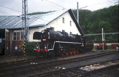A stationary, black steam locomotive rests on tracks beside a functional rail yard building with a metal roof and visible ele...