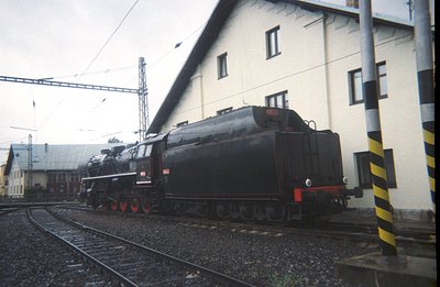 A weathered, stationary steam locomotive rests on tracks beside a plain, industrial building. The locomotive exhibits dark gr...