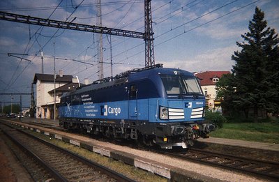A blue electric locomotive, likely a Siemens ES64U2, stands on a track in a rail yard. The locomotive displays "CargoNet" bra...