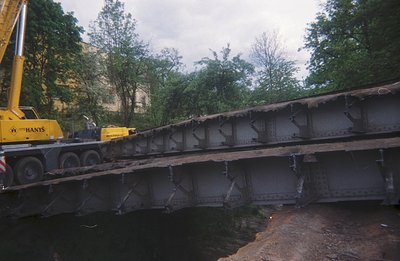 Large, weathered steel bridge section being transported, supported by a yellow crane. Visible details include bolted connecti...