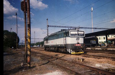 Detailed view of a green diesel locomotive, possibly a class 66, on a rail line. Background shows a station platform, greener...