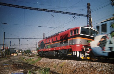 A red and grey electric locomotive, likely a BD series, moves along a track with overhead lines, typical of Bulgarian railway...