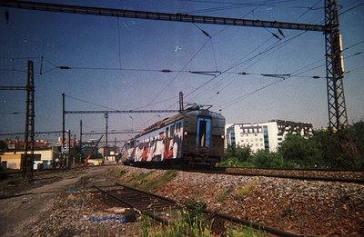A graffiti-covered electric train passes along a track, framed by overhead wires and utility poles. Visible are residential b...