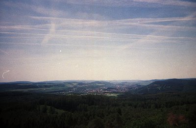 Elevated view of a valley town nestled amongst forested hills. Prominent contrail patterns dominate the sky, hinting at aeria...
