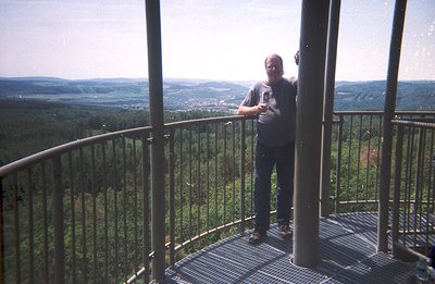 A man stands on a circular metal observation platform, overlooking a sprawling forested valley and distant town. He wears a d...