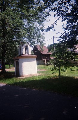 A small, whitewashed shrine sits beside a paved road, framed by lush foliage and a traditional wooden house. The shrine featu...