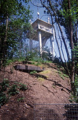 A tall, concrete observation tower rises above a wooded hillside. The tower features a circular platform and multiple antenna...