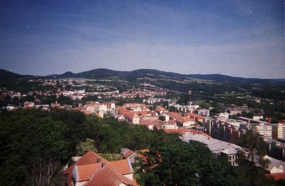 A wide, elevated view of a town nestled within a hilly, forested landscape. Predominantly red-tiled roofs characterize the ar...