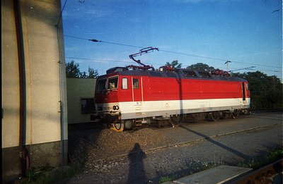 Striking, brightly colored electric locomotive stands on tracks with overhead power lines visible. Red and white livery indic...
