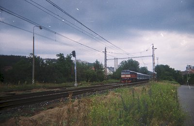 A commuter train travels on tracks alongside foliage & a platform. Overhead wires and pylons are visible against a cloudy sky...