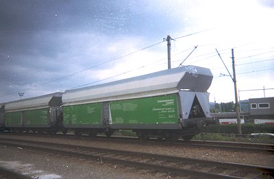 A long, green freight rail car carries stacked slabs of what appears to be precast concrete, likely for construction. The car...