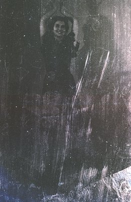 A grainy, black and white photograph shows a woman appearing to dance or gesture joyfully. Visible wear suggests age and hand...