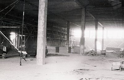 An industrial interior shows stacked lumber, supporting pillars, and a complex system of scaffolding. Appears to be an unfini...