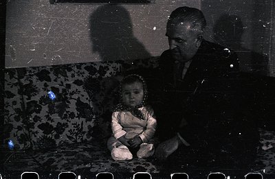 A seated man, in a suit, leans toward a young child wearing a patterned dress and a bow. They are positioned on a floral-patt...