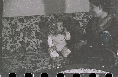 A baby wearing a patterned bonnet sits on a floral-upholstered sofa, gesturing towards a woman seated nearby. Likely a candid...
