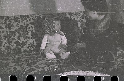 A seated toddler, clad in a romper and headscarf, is held by an adult, likely a parent. Detailed floral upholstery is visible...
