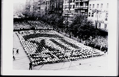 Crowd forms a large, grid-like pattern on a paved plaza. Tall buildings frame the scene, with a signage visible at the upper ...