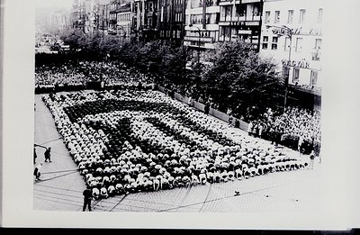A massive crowd forms a human shape, seemingly a stylized figure or emblem, occupying a large plaza surrounded by urban build...