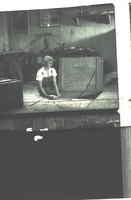 A figure in work overalls kneels, seemingly examining a floor surface within an interior space with visible structural beams ...