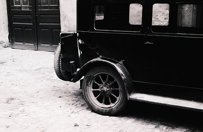 A vintage, black sedan with wire-spoke wheels sits on a cobbled street. The car’s side panel and a portion of the rear door a...