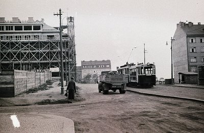 Construction site & tram meet on a street. A man walks past a truck, with an older-style tram in the background. Building sca...