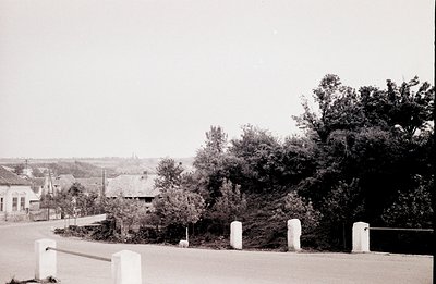 A roadside scene captures a small town with traditional architecture. A stone barrier lines the foreground, framing a view of...